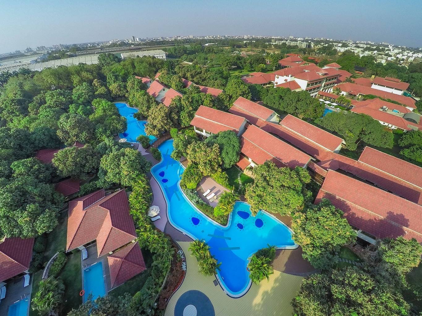 Aerial view of Madhubhan Resort & Spa: red-tiled villas and a winding pool among mature trees, the city beyond